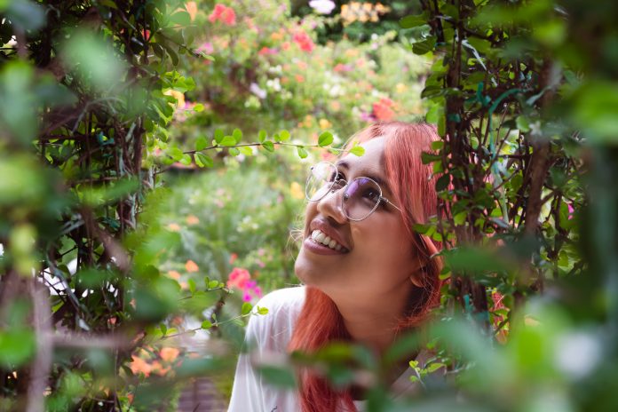 Diverse Asian woman with red hair looking up at colourful flowers in Botanical garden - View of young happy millennial girl smiling outdoors through a rose bush - Nature, weekend & lifestyle concept