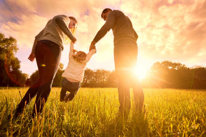 Glad familj i parkens kvällsljus. Ljusen av en sol. Mamma, pappa och baby glad promenad i solnedgången. Konceptet av en lycklig familj.Föräldrarna håller barnets händer.