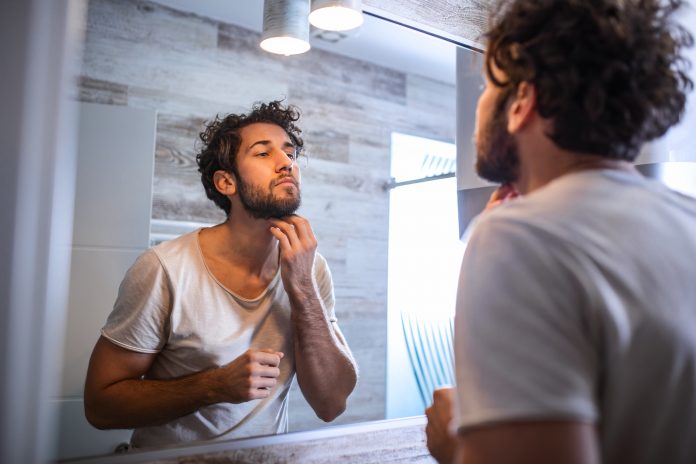 Reflection of handsome man with beard looking at mirror and touching face in bathroom grooming