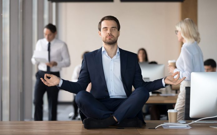 Mindful calm businessman in suit meditating at office sitting in lotus position on work desk, successful ceo executive doing yoga exercise at workplace, peace of mind, no stress free relief concept