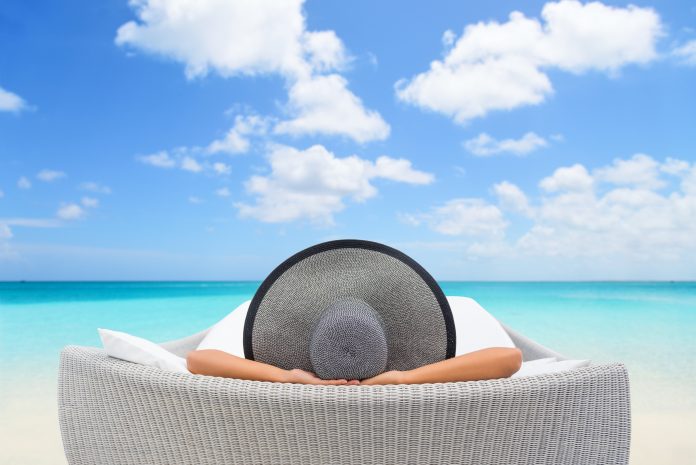 Travel vacation woman relaxing lying down on sun bed sofa lounge chair on holidays. Sleeping person lounging in hat on outdoor beach daybed lounger on ocean background luxury vacation.