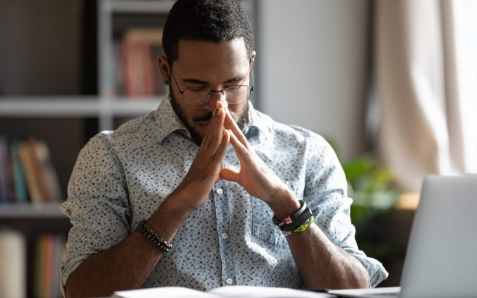 Young african american ethnic businessman praying asking for help concerned about problem making difficult decision concentrating mind meditating put hands in prayer sit at work desk in office