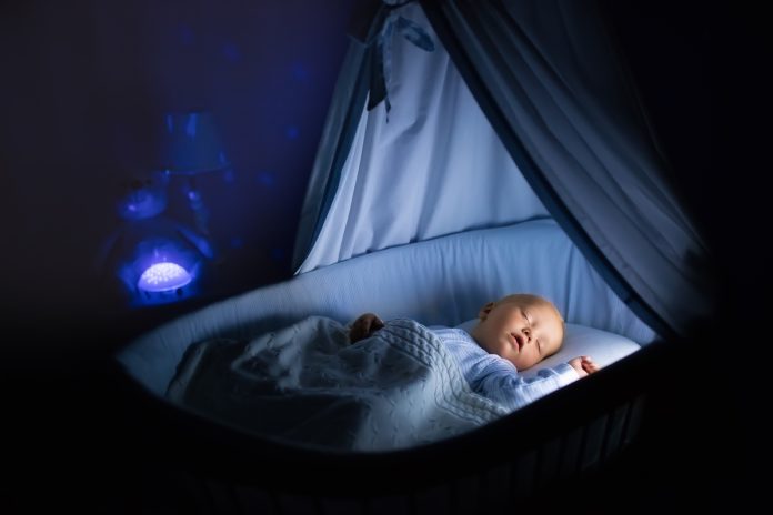 Adorable baby drinking milk in blue bassinet with canopy at night. Little boy in pajamas with formula bottle getting ready to sleep in dark room with crib, lamp and toy bear. Bed time drink for kids.