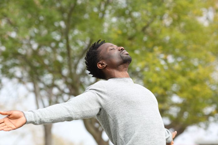 Schwarzer Mann atmet frische Luft und streckt die Arme in einem Park mit einem grünen Baum im Hintergrund aus