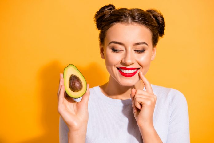 Close up photo beautiful she her lady pretty hairdo hold slice avocado look side with interest hand arm finger chin yummy tasty wear casual white pullover clothes isolated yellow background.