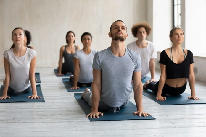 Diverse people wearing sportswear doing cobra exercise at group lesson, practicing yoga on mats, stretching in Bhujangasana pose, working out in modern yoga studio with young male instructor