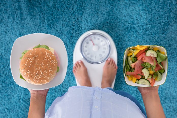 High Angle View Of A Person Holding Burger And Salad Standing On Weighing Machine