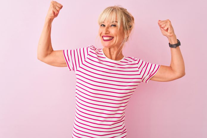 Mittlere Alter Frau trägt gestreiftes T-Shirt stehend über isolierten rosa Hintergrund zeigt Arme Muskeln lächelnd stolz. Fitness-Konzept.