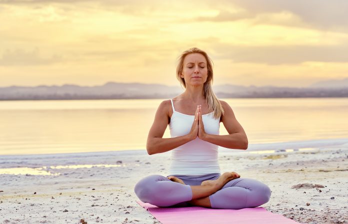 Middle aged woman in activewear make namaste gesture sitting in lotus position on beach near sea during sunset or sunrise closed eyes do meditation on nature, balance harmony healthy lifestyle concept