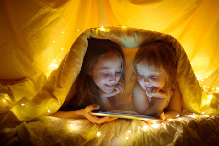 Two kids using tablet pc under blanket at night.