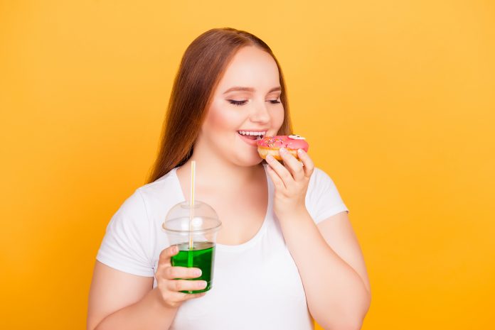 Bodycare body care concept. I don't want to be on a diet! Excited cheerful joyful plump woman, white tshirt, eating tasty glazed pink donut enjoying soft drink with a straw, isolated on background