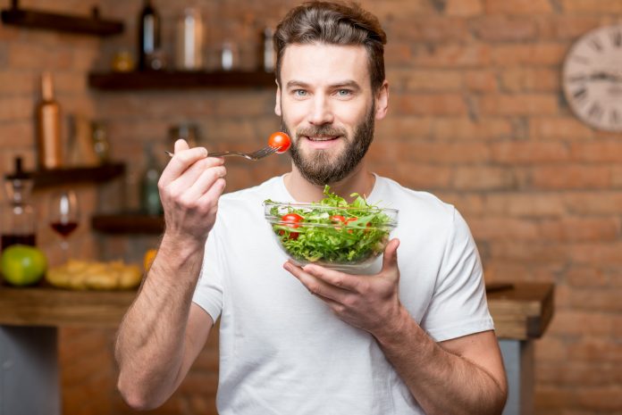 Uomo bello e barbuto in maglietta bianca che mangia insalata con pomodori in cucina. Concetto di cibo sano e vegano