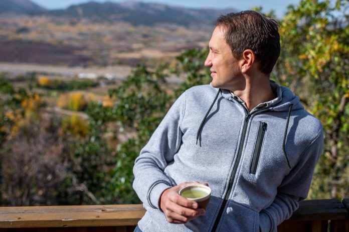 Happy man standing in backyard garden at home drinking tea with view of rocky mountains in Aspen, Colorado during autumn