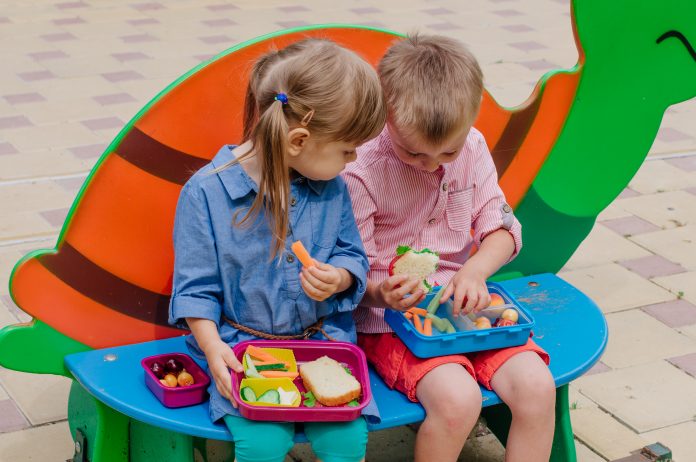 Healthy food concept. Girl and boy preschool students eating their lunches of sandwich, fresh vegetables and berries from lunch boxes sitting outdoor.