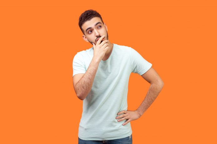 Need to think. Portrait of thoughtful brunette man with beard in white t-shirt holding his chin and pondering idea, confused not sure about solution. indoor studio shot isolated on orange background
