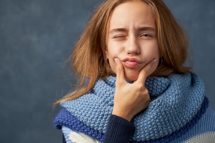 Portrait happy female laughs at something funny, has positive expression. Teenage girl with long thick blonde hair wearing warm woolen blue sweater and scarf is isolated against texture wall in Studio