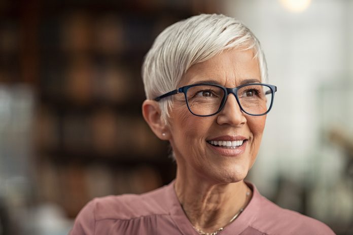 Portrait of smiling mature woman with spectacles and short grey hair in library looking away. Senior librarian standing in reading hall and thinking. Old beautiful lecturer contemplating in library. Future and vision concept.