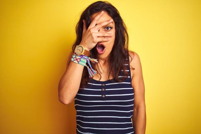 Young beautiful woman wearing striped t-shirt standing over isolated yellow background peeking in shock covering face and eyes with hand, looking through fingers with embarrassed expression.