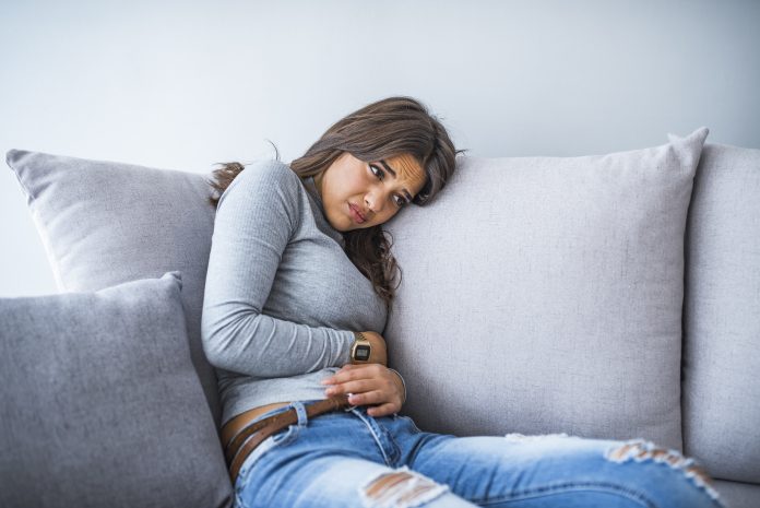Young woman suffering from abdominal pain while sitting on bed at home. Young woman suffering from abdominal pain at home. Gynecology concept. Young woman in pain lying on couch at home, casual style indoor shoot