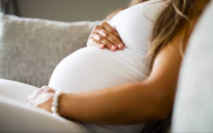 Primo piano di una donna incinta che si rilassa e si siede di lato sul divano. Si tiene le mani sulla pancia.