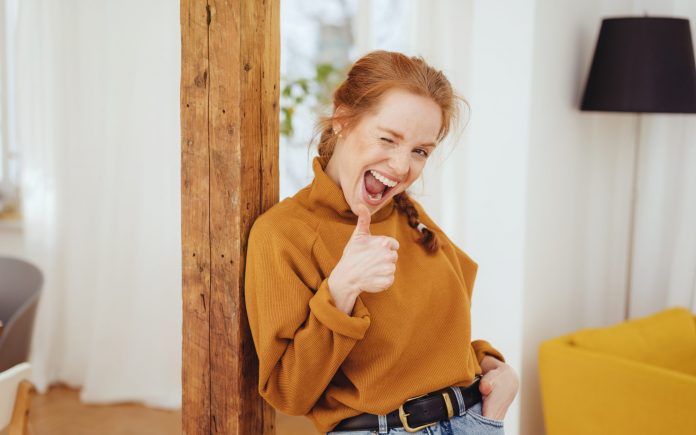 Cute playful young woman winking and giving a thumbs up gesture of success or agreement as she leans against a wooden pole indoors