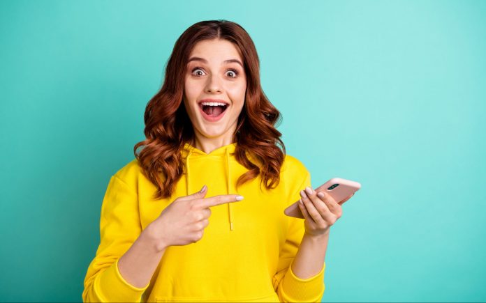 Photo of beautiful cheerful ecstatic attractive curly wavy trendy stylish, youngster pointing at telephone held with hands wearing yellow sweater hoodie isolated over vivid teal color background