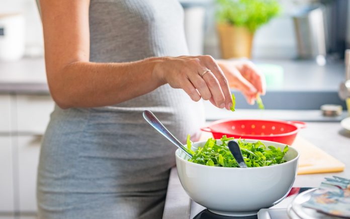 Femme enceinte se lavant les mains et choisissant une salade dans la cuisine ; saladier blanc et saladier rouge à l'arrière.