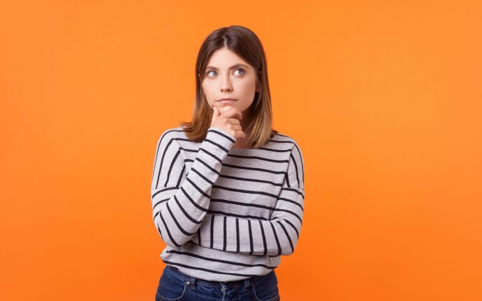 Think up plan. Portrait of pensive woman with brown hair in long sleeve striped shirt standing holding hand on chin and looking up, thinking intensely. indoor studio shot isolated on orange background