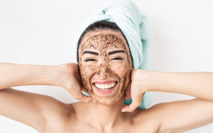 Young smiling woman applying coffee scrub mask on face - Happy girl having skin care spa day at home - Healthy alternative natural exfoliation treatment and people lifestyle concept