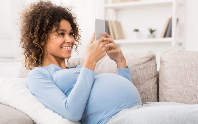 Afrikanisch-amerikanische schwangere Frau beim Surfen auf dem Smartphone, zu Hause auf dem Sofa liegend, Freiraum