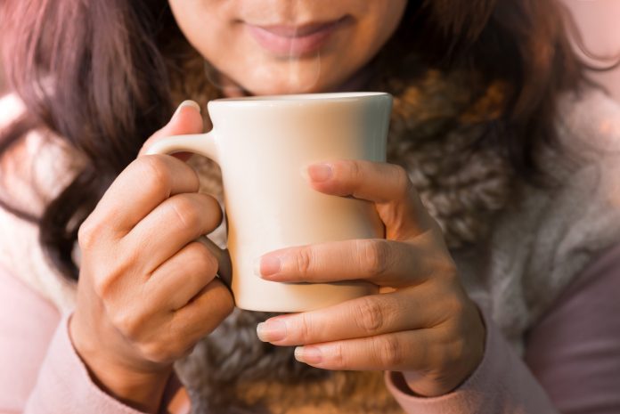 Nahaufnahme von Frau Hände halten eine Tasse Kaffee.  Weiblich in Wintermode Kleidung genießen heißes Getränk mit lächelndem Gesicht, selektiv fokussiert.