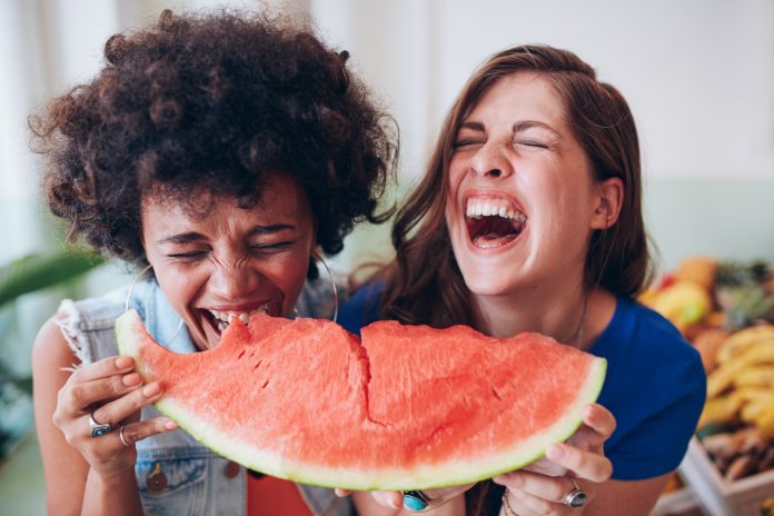 Портрет крупным планом двух молодых девушек, наслаждающихся арбузом. Подруги едят арбузную дольку и смеются вместе.