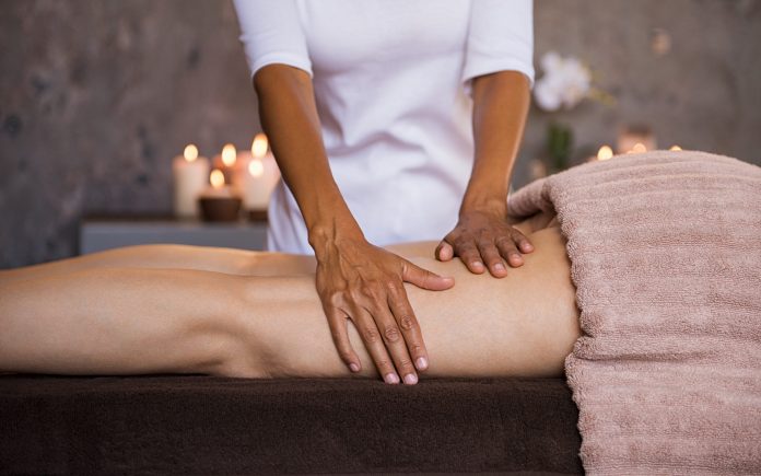 Therapeutic massage for senior woman by beautician at spa salon. Closeup of professional masseuse hands massaging woman legs at wellness center. Anti cellulite treatment and leg drainage on mature woman.