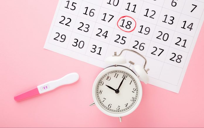 Calendar, test with two stripes and white alarm clock on pastel pink table background. Positive result. Pregnancy time beginning. Closeup. Top down view.