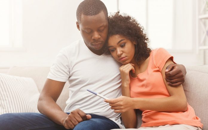 Surullinen afrikkalais-amerikkalainen pari negatiivisen raskaustestin tuloksen jälkeen, istuu sohvalla kotona, kopioi tilaa