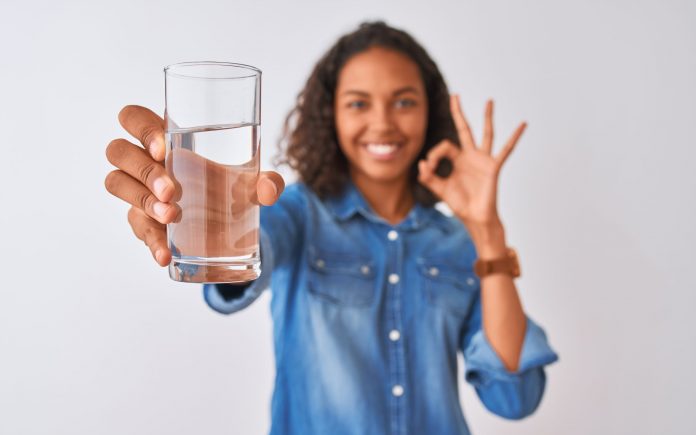Young brazilian woman holding glass of water standing over isolated white background doing ok sign with fingers, excellent symbol