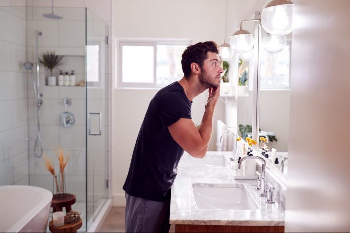 Mann im Pyjama steht am Waschbecken und trägt Feuchtigkeitscreme im Badezimmer auf